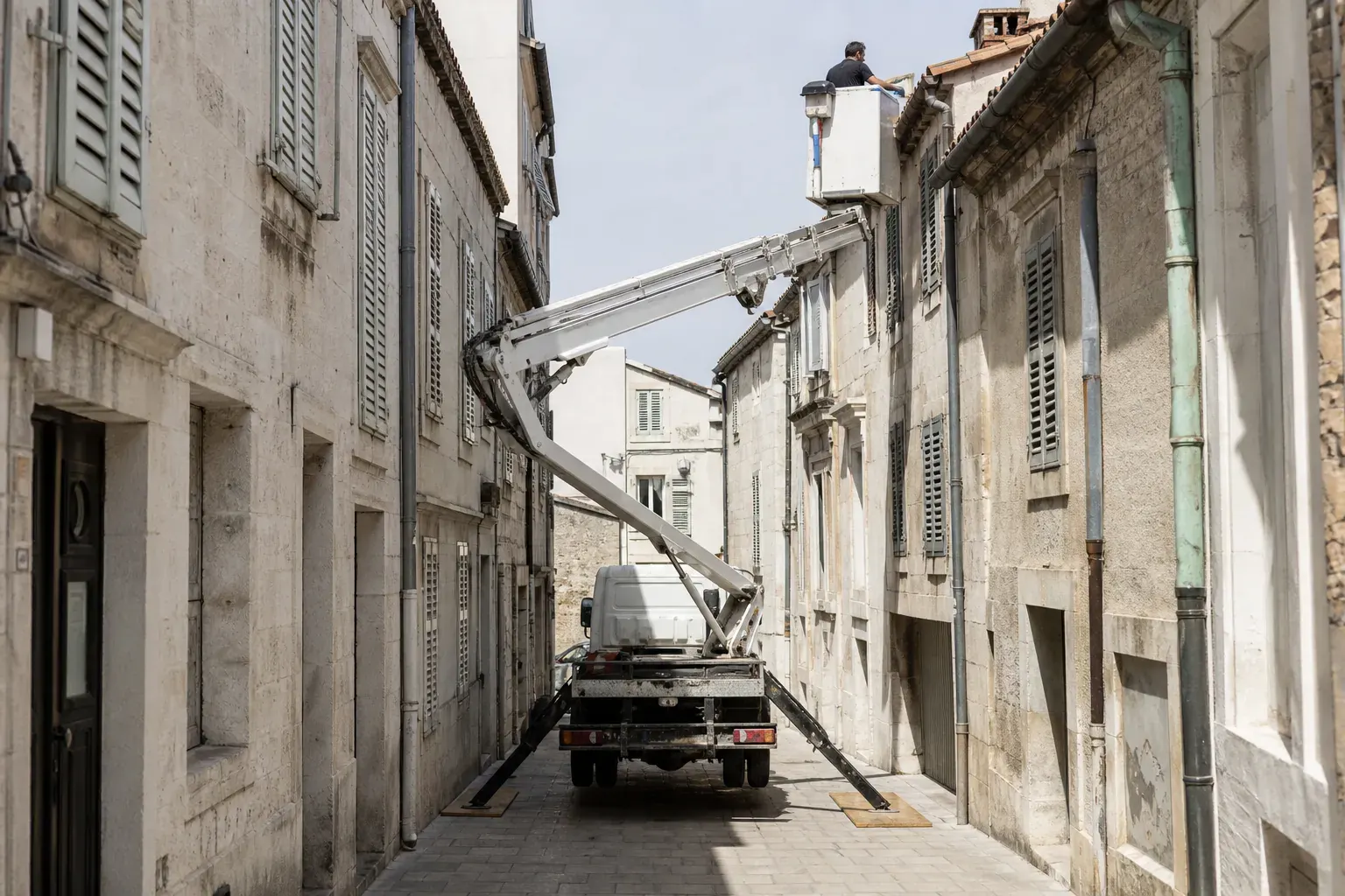 Nacelle sur camion calée dans une rue étroite de centre ancien, panier levé vers l’égout d’un toit au troisième étage