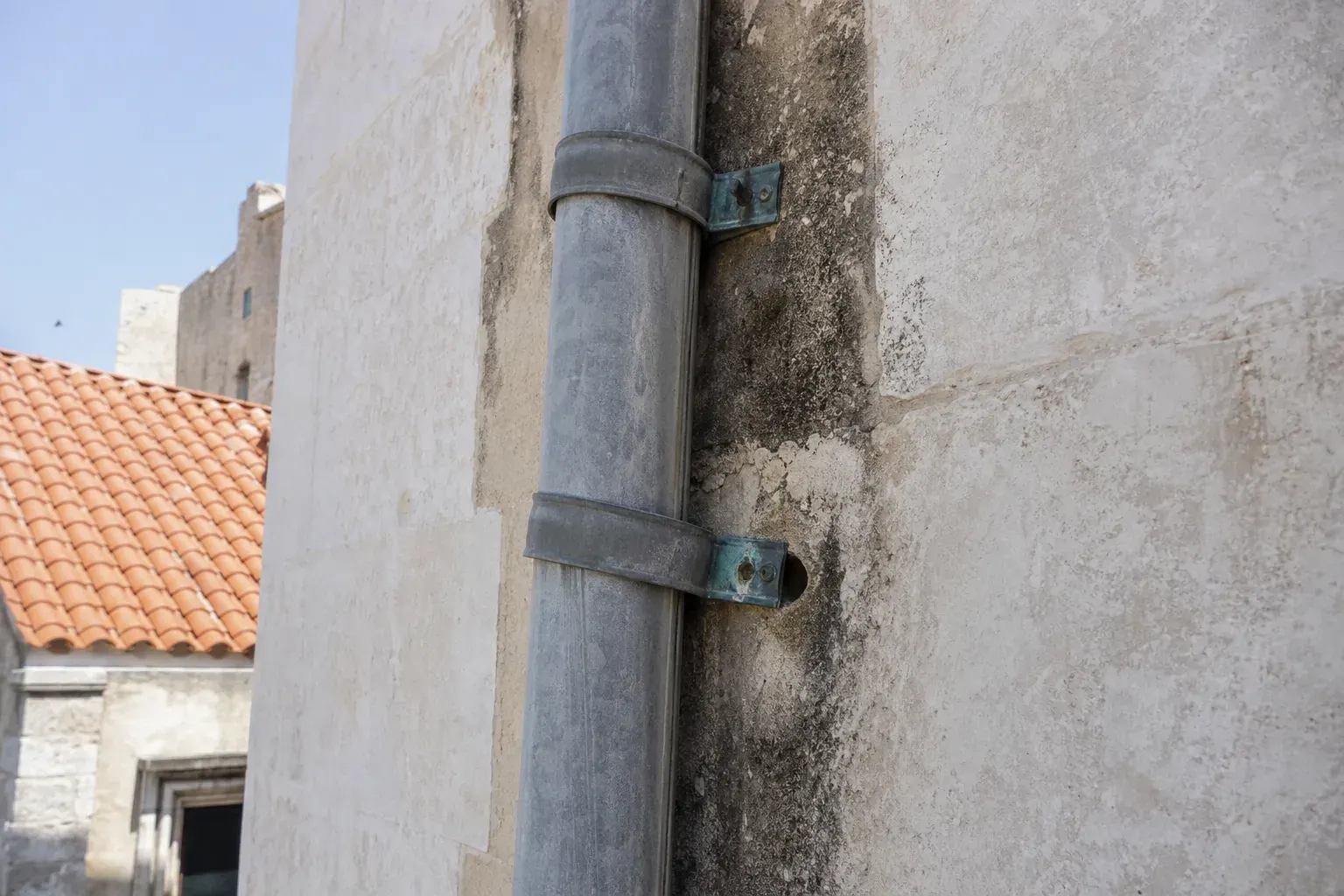 Gros plan sur une descente d’eaux pluviales en zinc le long d’une façade claire, collier descellé et trace d’humidité sous le raccord